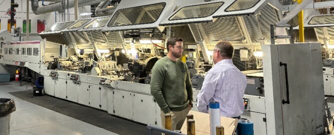 Two men standing next to some equipment in a factory - folding cartons